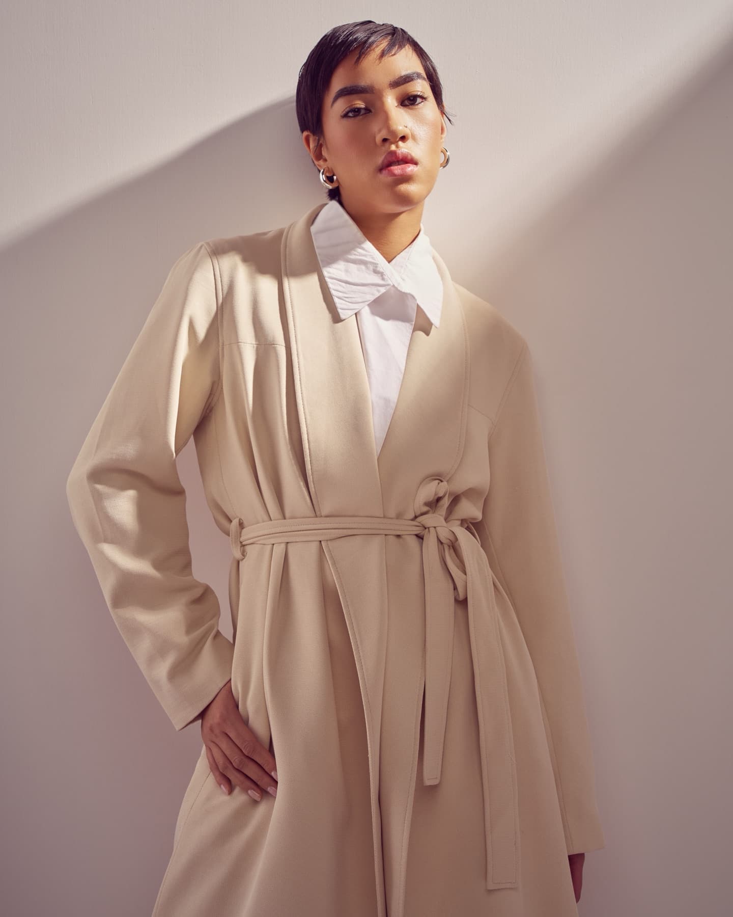 Model in a belted cream trench coat, lit by a hard slash of sunlight